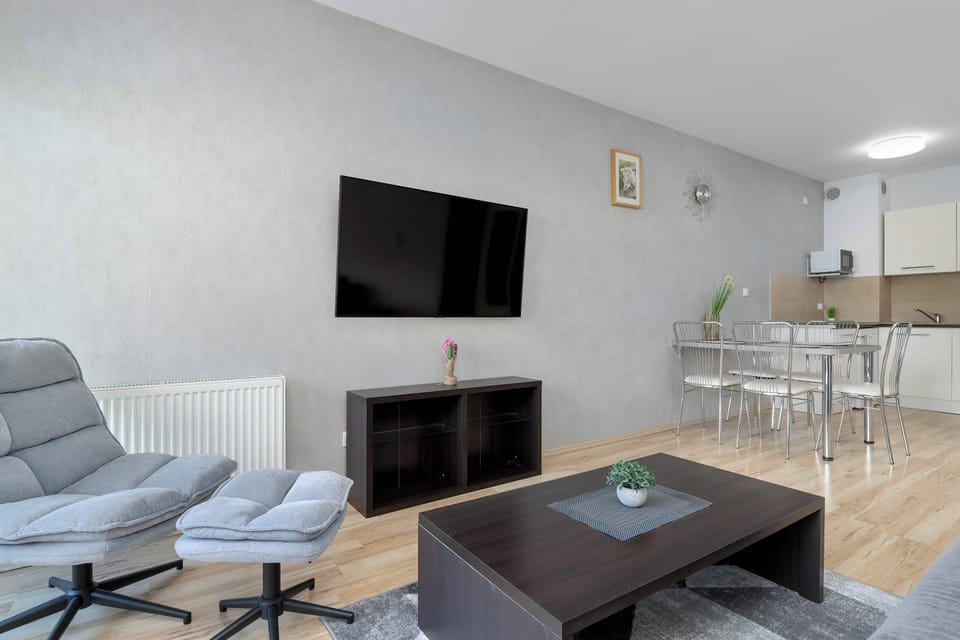 A wide shot of the studio shows a grey armchair with a footrest positioned near the media area. The open layout connects the seating, dining, and cooking zones seamlessly.