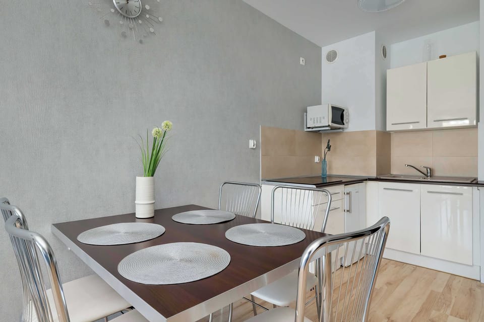 The dining table is shown from a low angle, highlighting the place settings and the white kitchenette in the background. A vase with green plants adds a fresh touch to the room's decor.