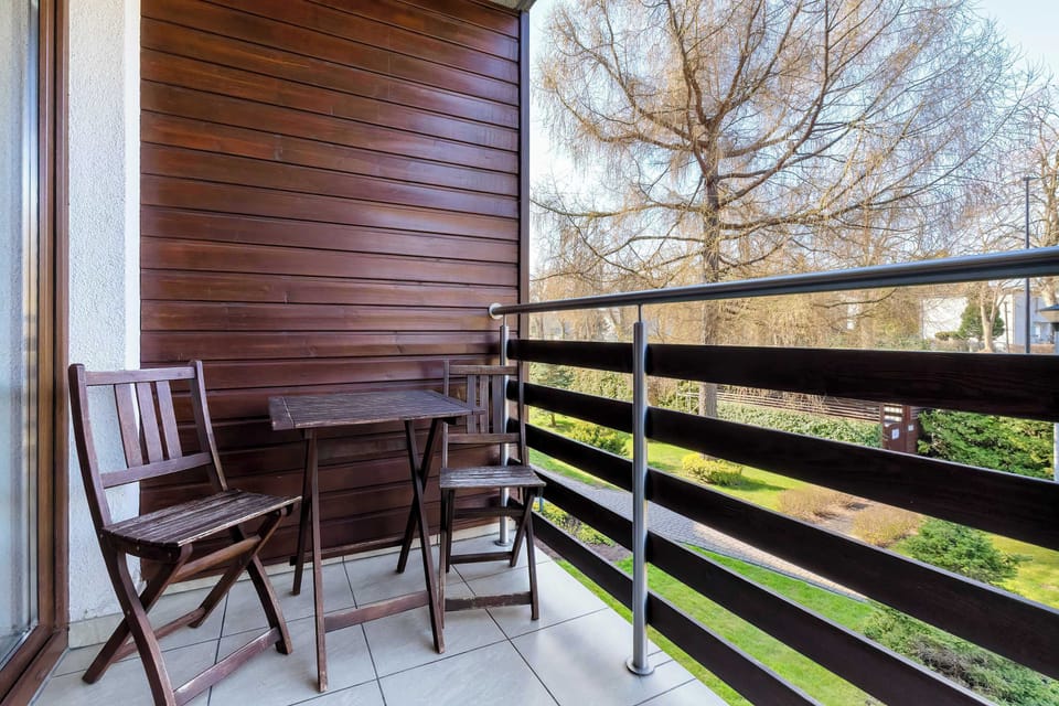The private balcony is furnished with a wooden bistro set, including a small table and two folding chairs. It overlooks a green area with mature trees and a manicured lawn.