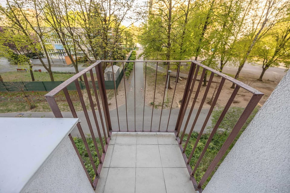 A modest balcony with a simple metal railing overlooks a peaceful, leafy courtyard filled with mature green trees.