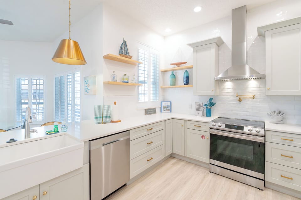 White shaker kitchen with farmhouse sink, gold pendant lighting, open shelving, and stainless appliances — stylish vacation rental kitchen in Palm Coast, FL.