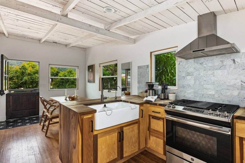 The full kitchen — farmhouse sink, gas range, and wood-slab counters under whitewashed beams.