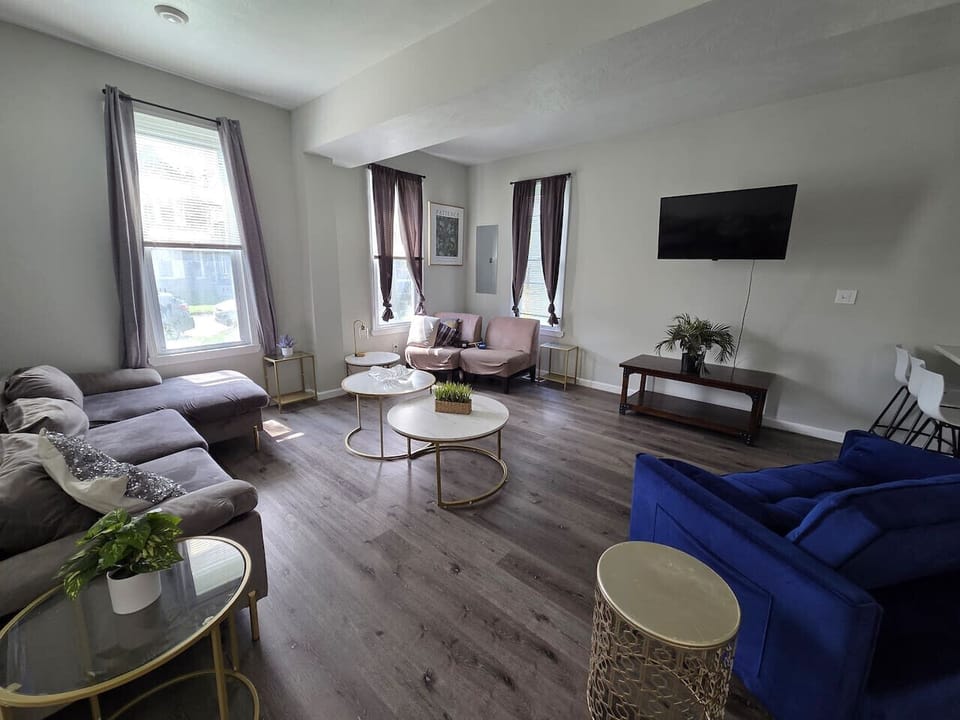 This bright and airy open-concept living area seamlessly connects to the kitchen, featuring sleek grey wood-toned flooring and modern recessed lighting throughout. The space offers ample seating with a plush velvet sectional and a striking cobalt blue lov