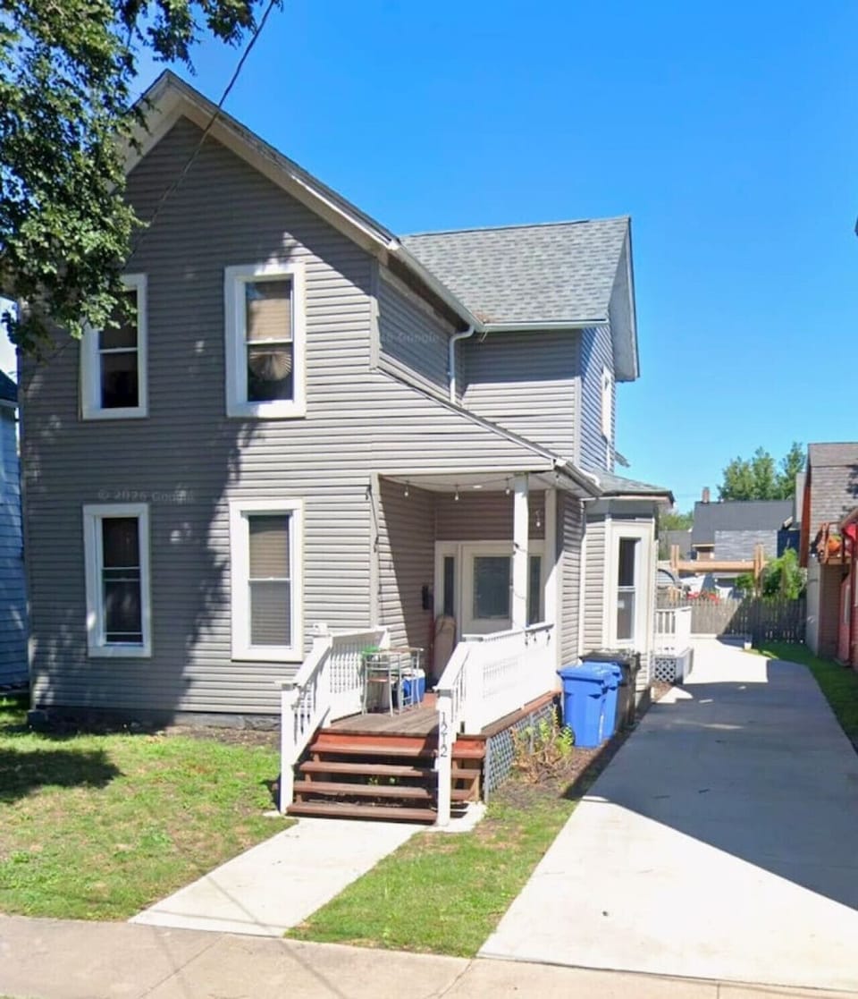 This charming two-story home features a classic exterior with clean grey siding and traditional white trim. A cozy front porch with white railings and wooden steps leads to the main entrance, creating a welcoming first impression for guests. The property 