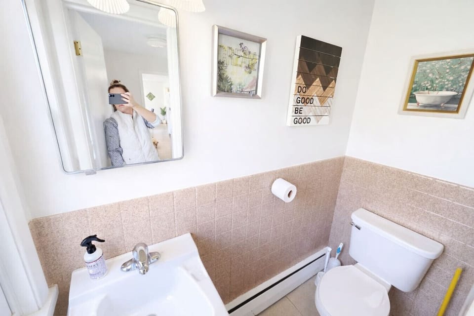 This bright and clean bathroom features timeless tan tiling and a large window draped in soft, airy linens for a refreshing start to your day. Bold floral accents and a spacious vanity create a charming, functional environment equipped with all the essent
