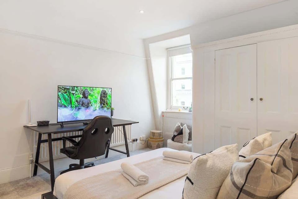 View the bedroom work layout showcasing a dark desk with a flat-screen TV monitor, ergonomic desk chair, white wardrobes, and window light.