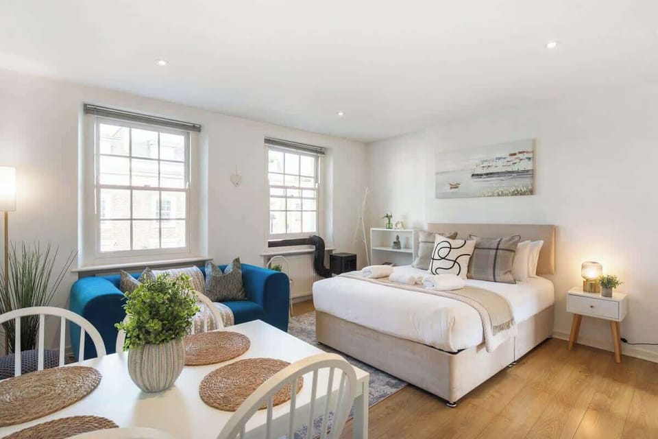 Relax in this bright, open-plan studio apartment featuring a dining table set in the foreground, a comfortable bed with stylish accent pillows, and a vibrant blue sofa situated beneath two large windows.