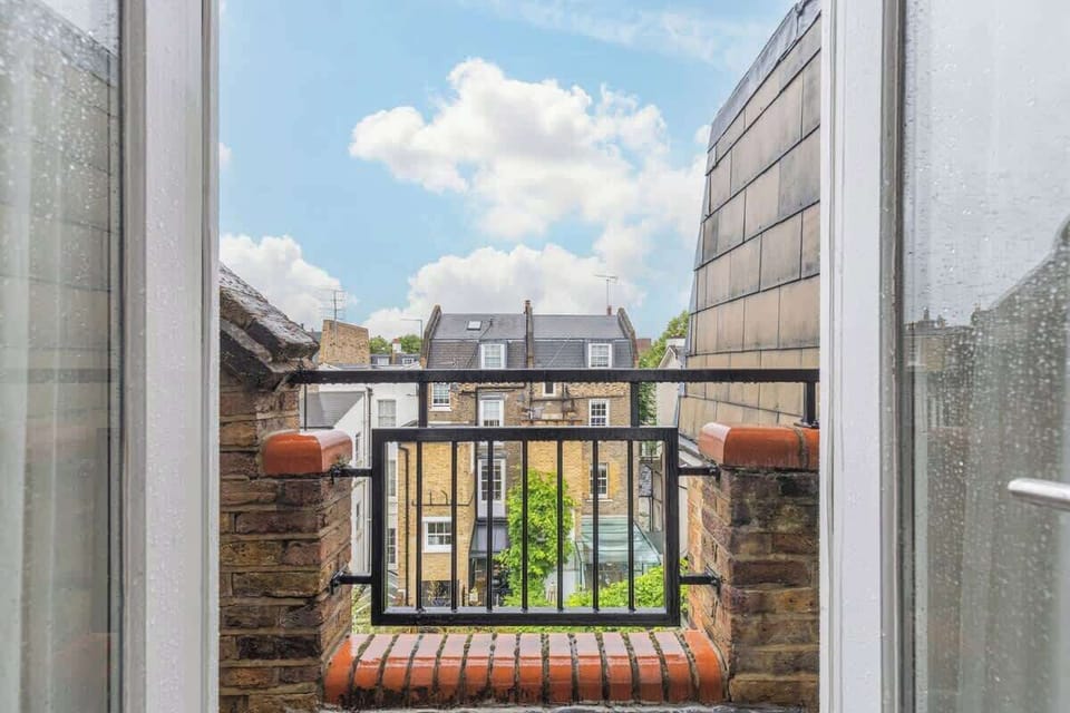 Step outside and enjoy the view from this cozy private balcony, framed by open doors and black metal railings looking out over surrounding neighborhood rooftops under a bright, cloud-filled sky.