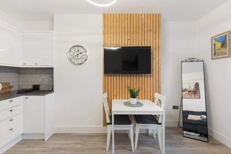 Gather for meals or watch TV in this bright dining space, featuring a white table with cross-back chairs, set against a modern wood-slat feature wall with a mounted flat-screen TV and adjacent white kitchen cabinetry.