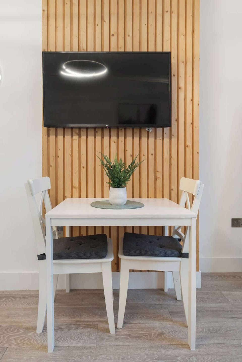 Dine or relax in this cozy nook featuring a white dining table with cross-back chairs, set against a modern light-wood slat accent wall with a mounted flat-screen TV.