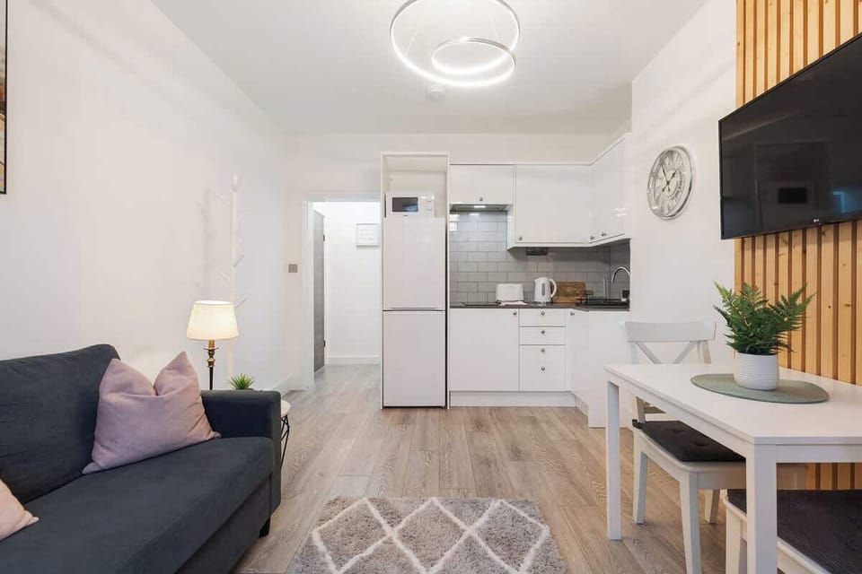 Take in the complete, beautifully layout of this open-concept apartment, featuring a plush charcoal sofa, a small white dining area, light wood-look flooring, and a fully equipped, sleek white kitchenette at the far end.