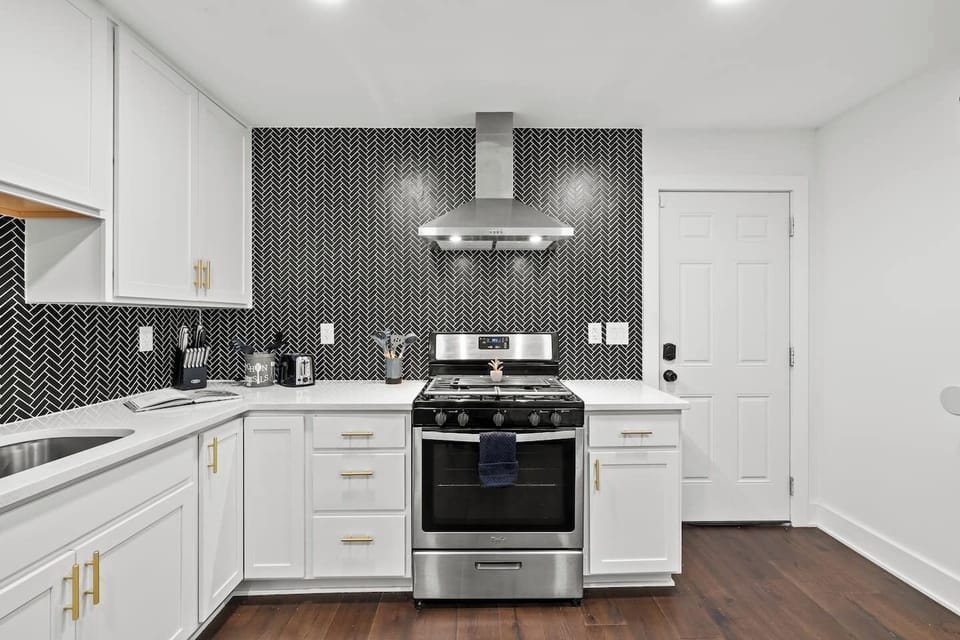 Sleek, fully equipped kitchen featuring white cabinetry, quartz counters, and gold accents. Highlights include a stainless steel stove/oven and a striking black herringbone backsplash. Perfect for preparing any meal.