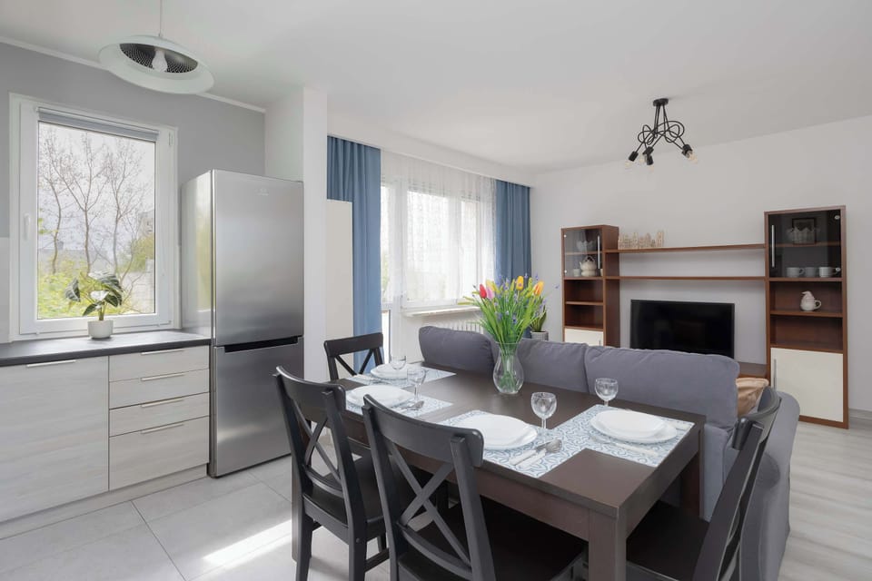 A dining area connected to a living space, featuring a dark table and chairs near a window. The room is clean and minimal, with soft tones and simple decor.