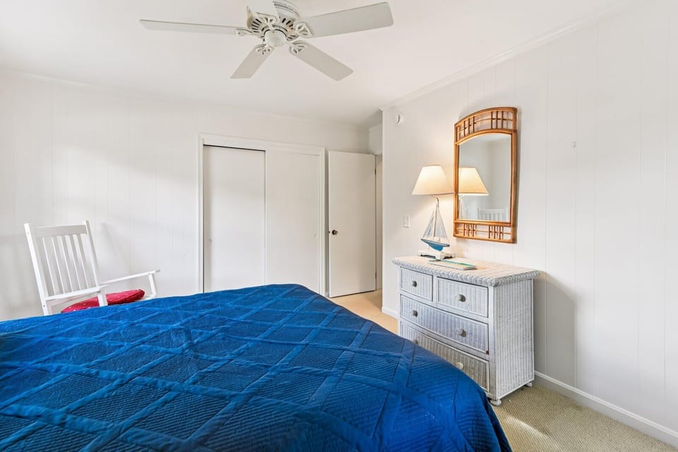 Your bright coastal bedroom awaits with a plush bed, breezy ceiling fan, charming nautical dresser, and cozy rocking chair by the window—perfect for unwinding after beach days.