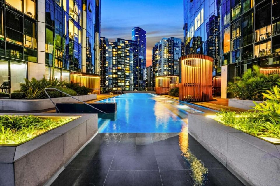 Rooftop lap pool set against a glowing city skyline at dusk, surrounded by lush greenery, warm timber cabanas, and soft landscape lighting — a serene urban retreat steps from your door.