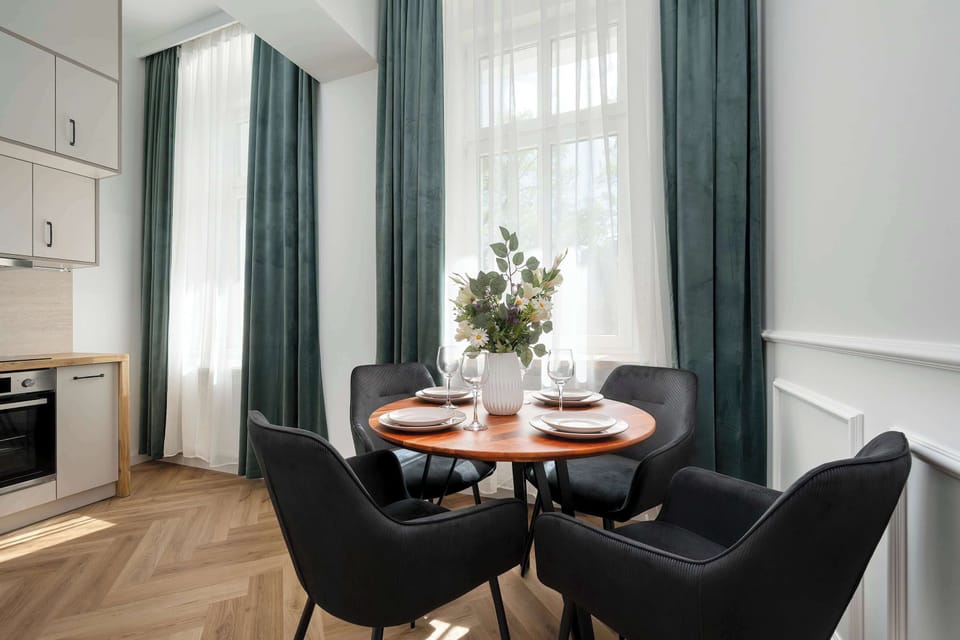 A dining area positioned by large windows with elegant curtains, featuring a round table and comfortable chairs. Natural light enhances the warm tones of the wooden floor and furniture.