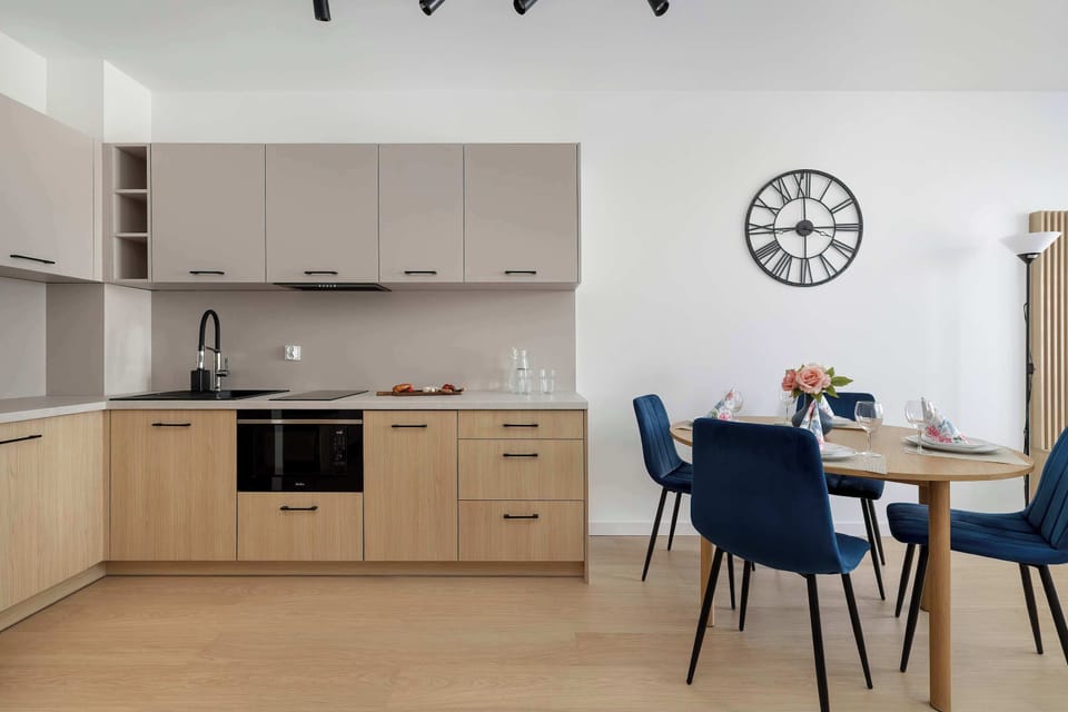 A compact kitchenette corner with light wood cabinetry and built-in appliances. A small dining setup with dark blue chairs sits next to it, complemented by a wall clock and simple wall decor.