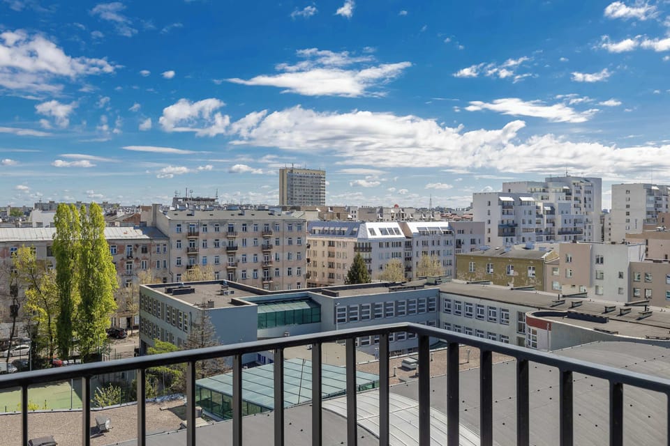 An expansive view from the balcony overlooks the rooftops and apartment buildings of the surrounding city. The horizon is filled with urban architecture under a bright, cloud-speckled sky.