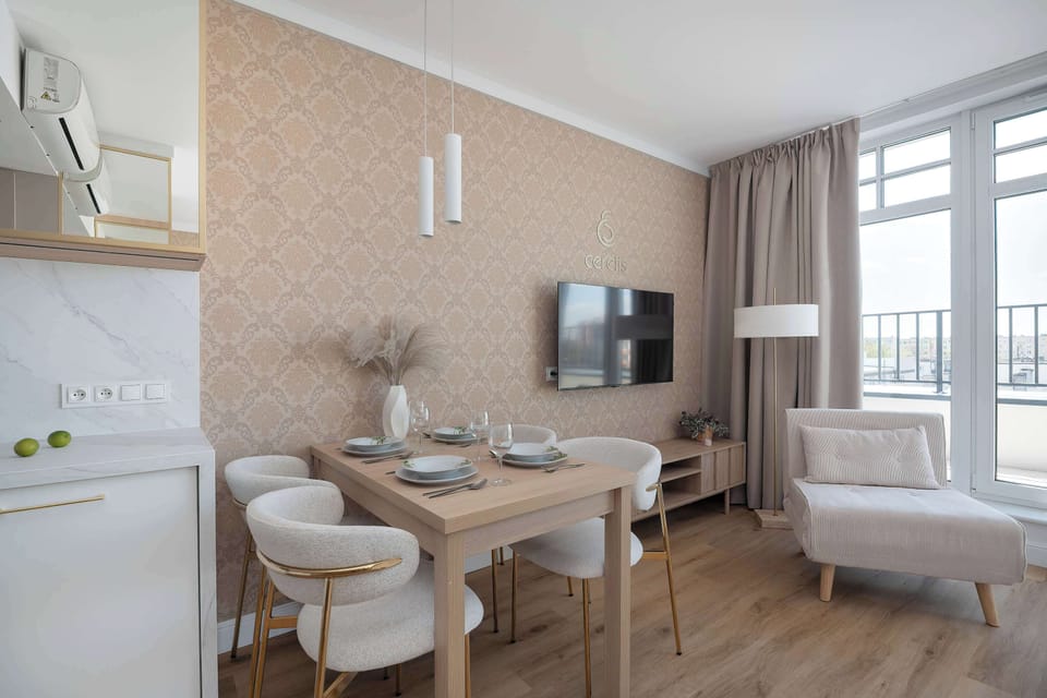 The dining area is shown next to a large window, featuring white chairs and a wooden table. A wall-mounted television and a white armchair are visible in the background lounge.