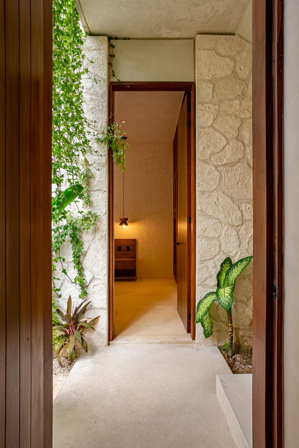 A serene hallway framed by stone walls and greenery, leading into a softly lit bedroom.