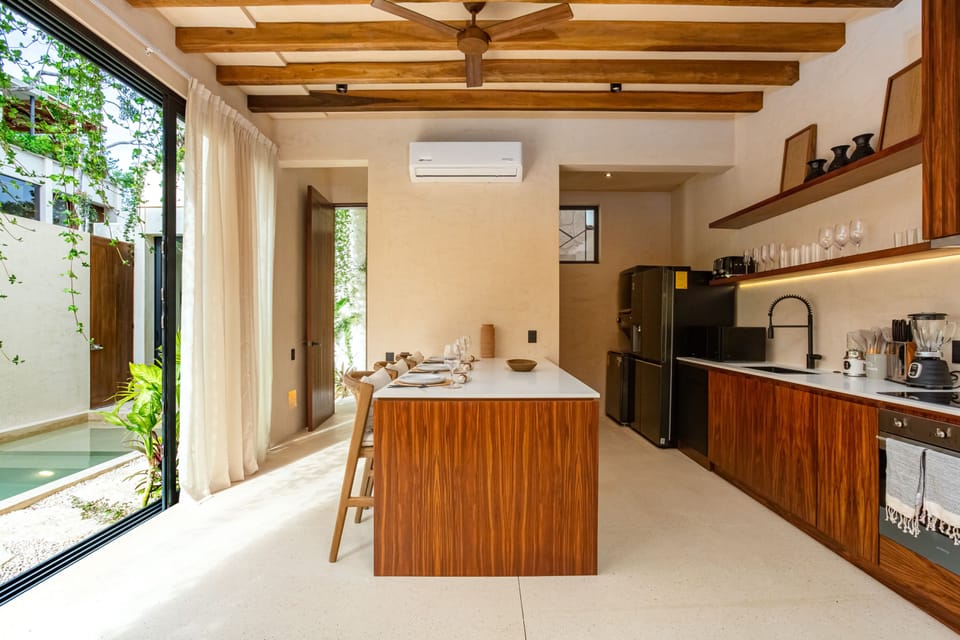 A bright, modern kitchen with wooden cabinetry, an island dining space, and large glass doors opening to a lush outdoor view.