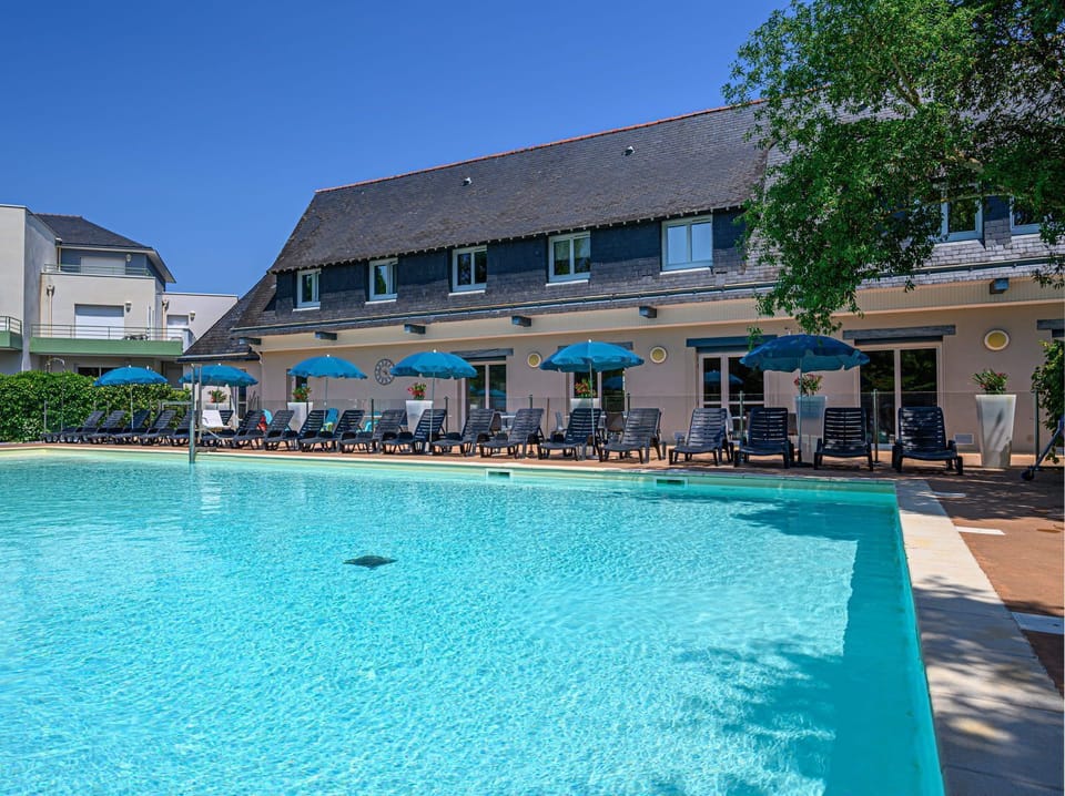 Outdoor heated pool with a row of dark loungers and blue umbrellas along the terrace — open May through September