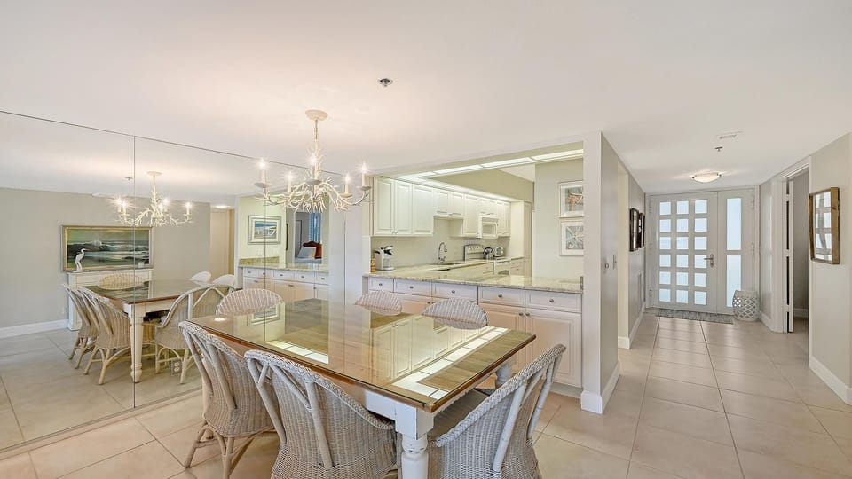 A glass-front wooden dining table surrounded by wicker chairs sits adjacent to a kitchen with granite countertops.