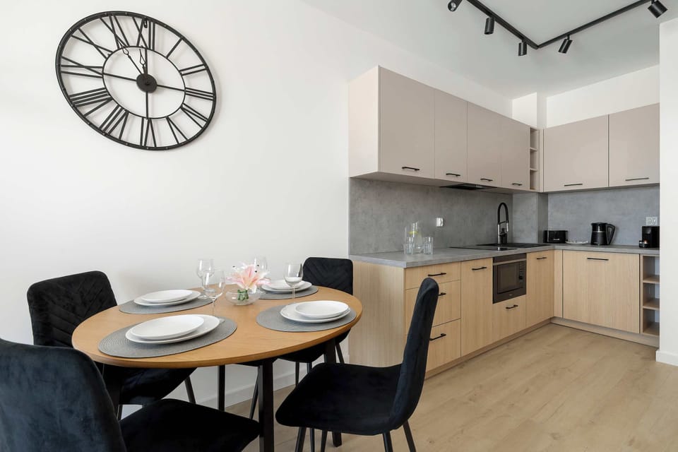 The dining area features a round wooden table and four navy blue chairs, set against a backdrop of a large wall clock. The kitchen and main living space are visible in the background.