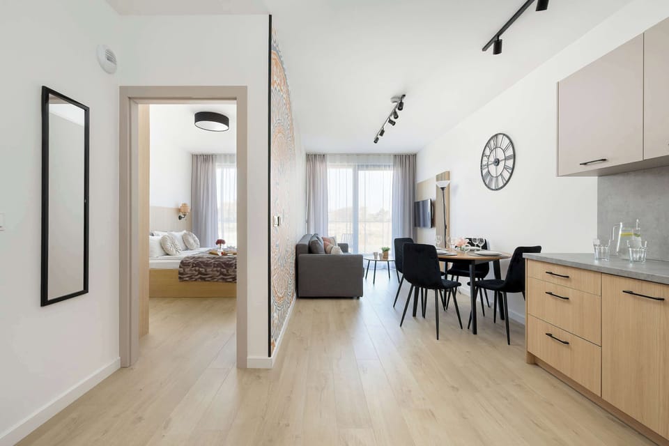 A view from the bedroom doorway looks out across the living area toward the bright balcony doors. The light wood flooring continues throughout the apartment, creating a seamless transition.