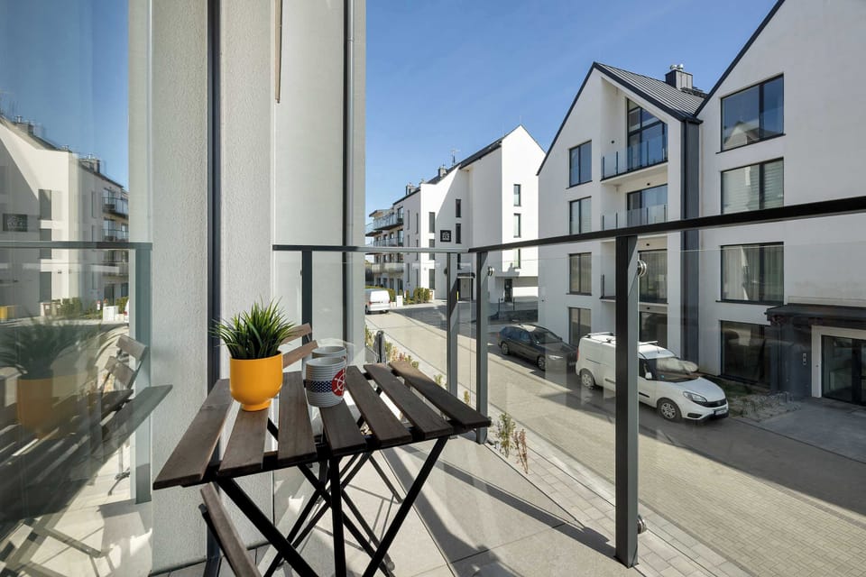 The balcony overlooks the street of a modern residential development with clean white buildings. A yellow flower pot on the table adds a cheerful touch to the outdoor seating.