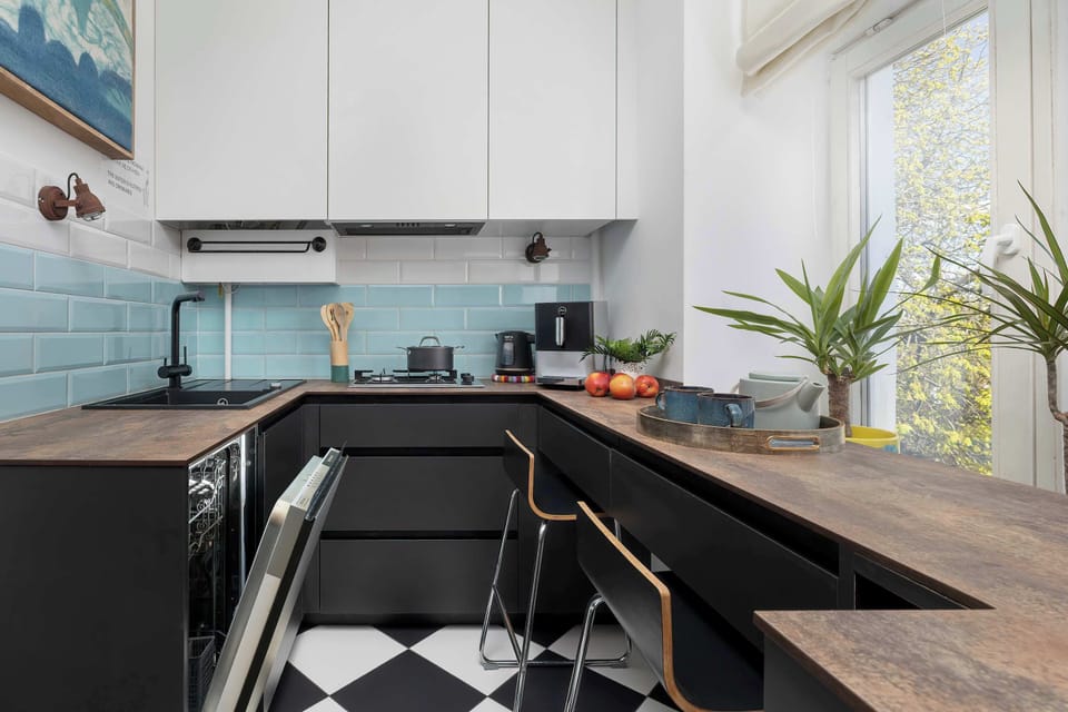 A view of the kitchen nook with a bar counter, next to which stand two metal bar stools, perfect for a quick breakfast. Green potted plants are visible in the background, bringing life to the minimalist space.