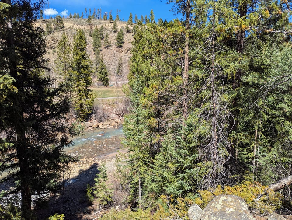 Pristine wilderness surrounds the property, featuring towering pine forests, rugged cliffs, and a crystal-clear river winding through the valley below.