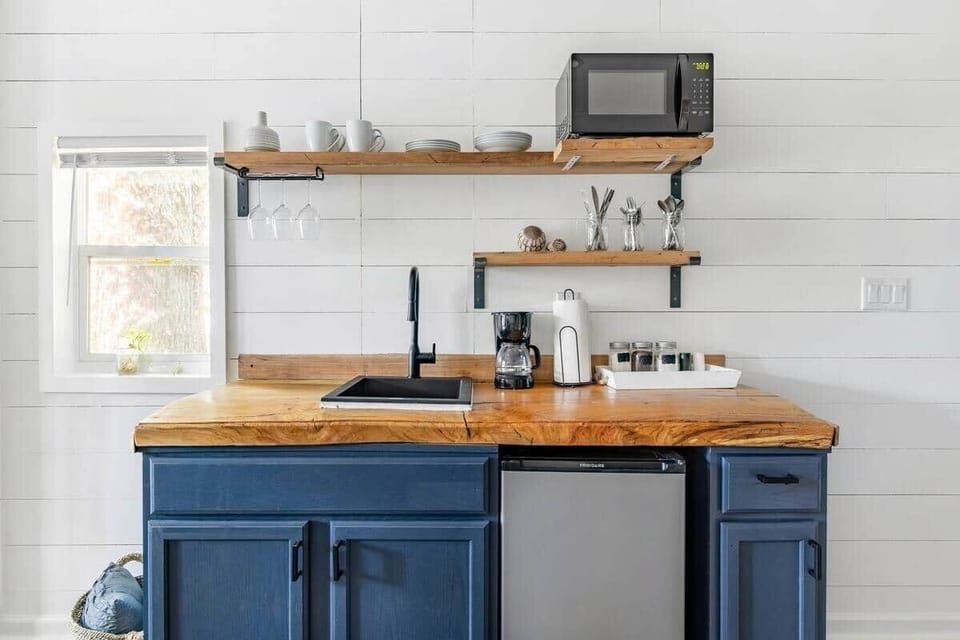 Bright kitchenette featuring deep blue cabinetry, natural wood countertops, and floating shelves with thoughtful essentials including a mini fridge, microwave, coffee maker, and modern matte black faucet.