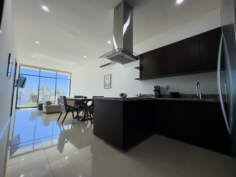 Black cabinetry and granite countertops define the kitchen, opening to a dining area with wood chairs and large windows.