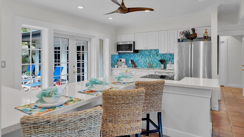 Gather around your fully equipped island kitchen with vibrant turquoise backsplash, modern appliances, and seating for casual meals while natural light streams through French doors opening to the pool area.