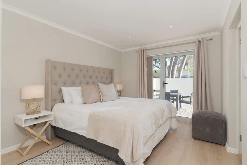 Bright main bedroom with tufted upholstered headboard, gold bedside lamp, and glass doors opening to garden views
