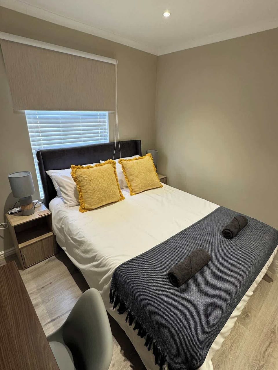 Bedroom with upholstered platform bed, mustard fringe cushions, timber nightstand and roller blind — cosy and neatly appointed