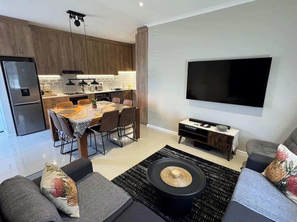 Bright open-plan living area — sectional sofa, large flat-screen TV, 8-seat dining table and fully fitted kitchen flowing together seamlessly.