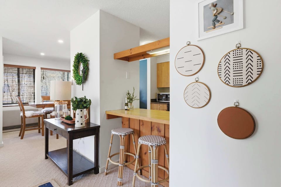 The dining area and kitchen pass-through from the entryway side — rattan bar stools at the counter and woven wall art give the space a relaxed, mountain-casual feel.