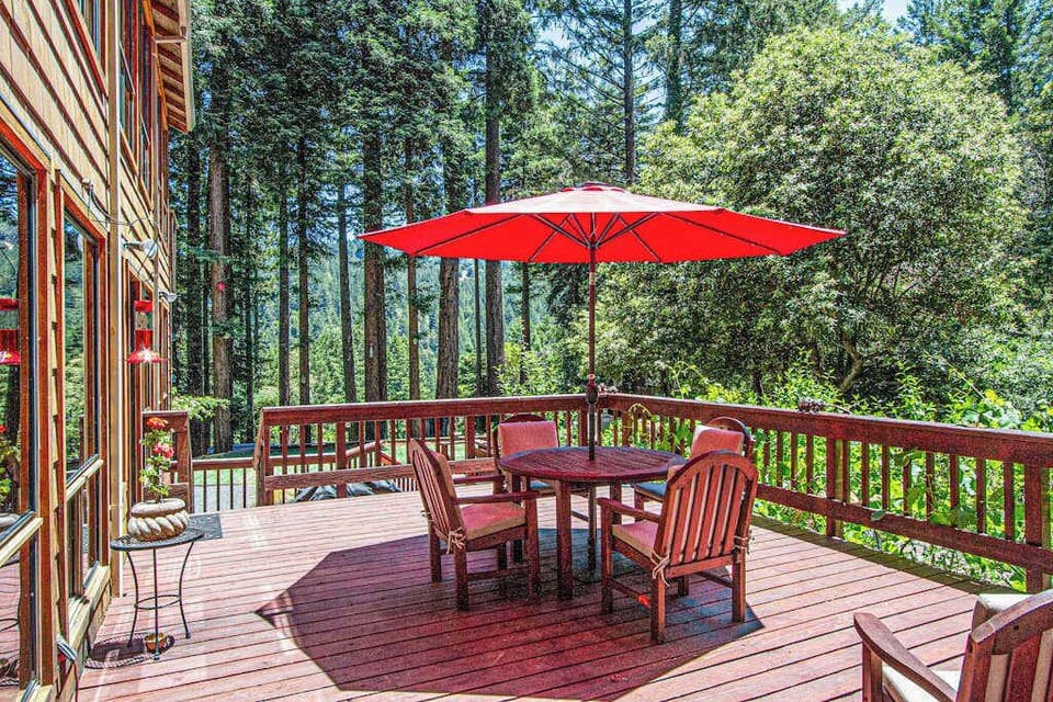 Savor your morning coffee on the spacious redwood deck — a shaded dining table for four awaits under a vibrant umbrella with forest canopy views all around.