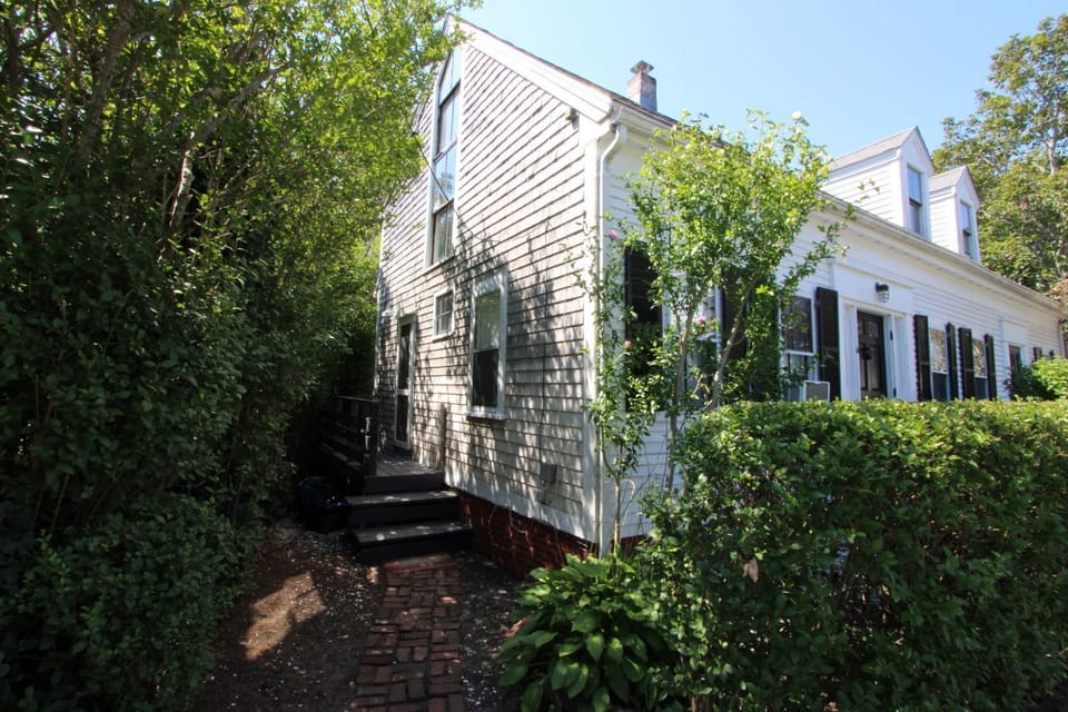 Charming shingled cottage nestled among trees and greenery, featuring classic black shutters and a welcoming brick walkway.