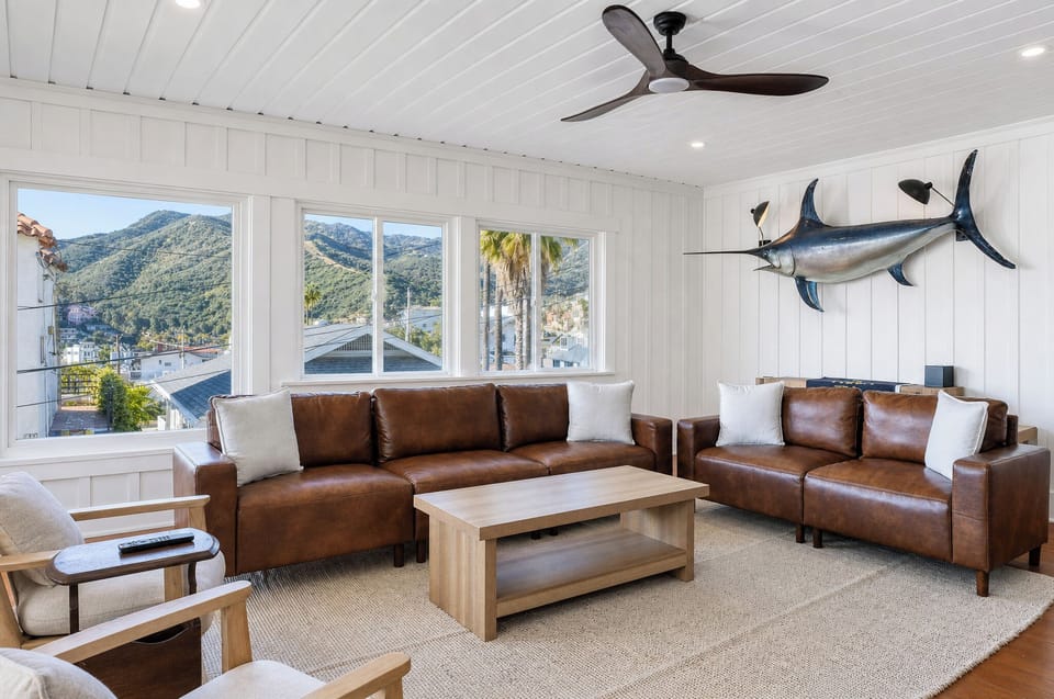 Relax on plush leather sofas beneath coastal decor and a ceiling fan, with mountain and palm tree views streaming through expansive windows inviting natural light.