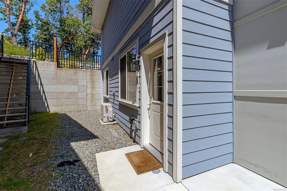 Side entrance with light blue horizontal siding, white door, concrete slab, and gravel ground beside a retaining wall.