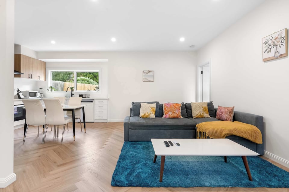 Unwind in this spacious living and dining layout, highlighting a grey sectional couch with vibrant cushions, a white coffee table, and an adjacent white dining set leading toward the kitchen.