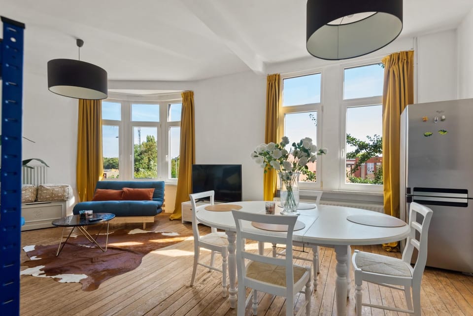 Sunny dining and living space with large windows and warm natural light. Enjoy meals at the bright dining table or unwind on the comfortable seating area while taking in open neighborhood views through the tall windows.