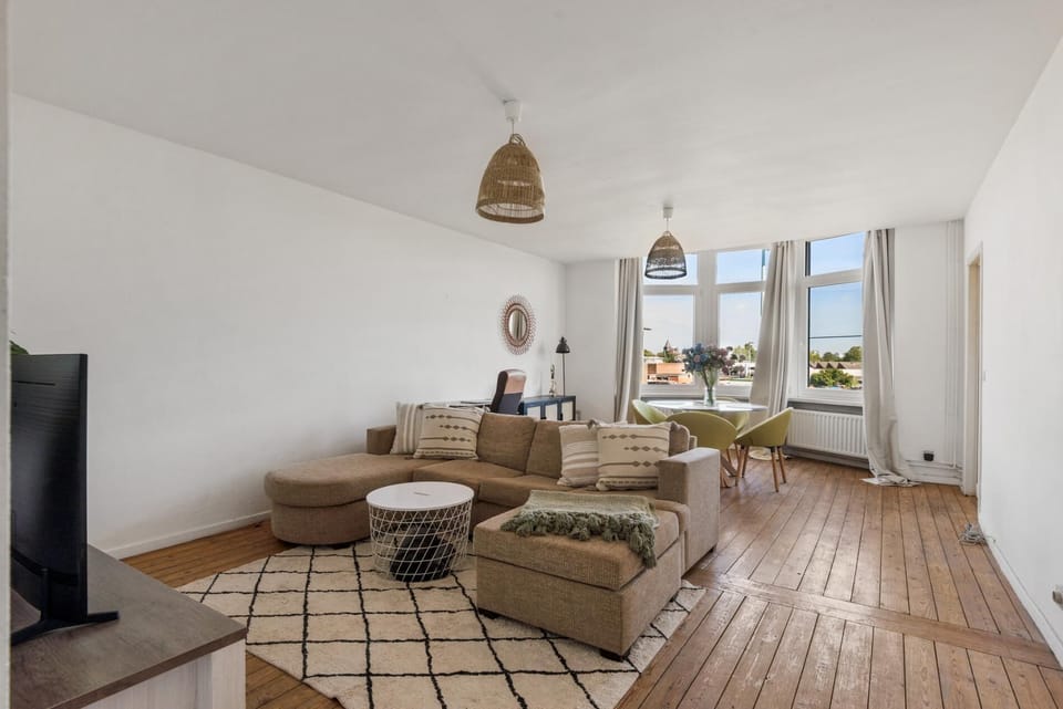 Spacious living and dining area with wooden floors, a comfortable sectional sofa, and large windows overlooking the neighborhood. The open layout offers plenty of natural light and a relaxed space for lounging, working, or sharing meals.