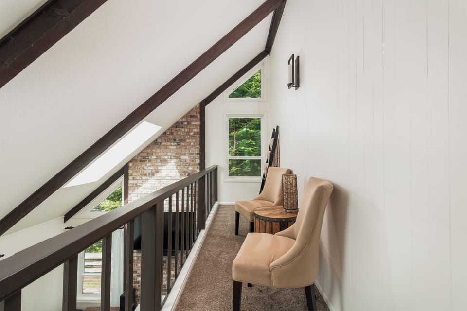 Relax in your private upstairs seating nook with vaulted ceilings, exposed wooden beams, and windows framing lush greenery—a peaceful retreat for morning coffee or quiet reading.