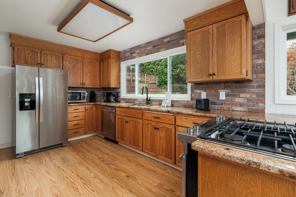 Your fully equipped kitchen features warm oak cabinetry, granite countertops, stainless steel appliances, and a gas range—perfect for preparing meals with views of the lush backyard through the bright windows.