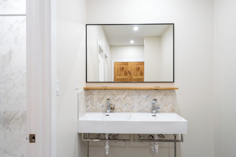 Sleek double vanity with herringbone tile backsplash and modern fixtures offers you a spa-like morning routine. Natural wood shelf and storage cabinets reflected above keep your essentials close at hand.