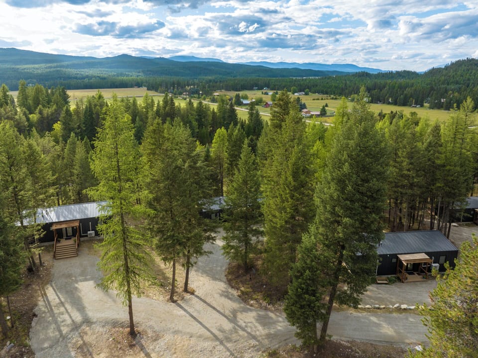 Aerial view showcases the property nestled among towering evergreens with mountain ranges creating a dramatic backdrop across the pristine wilderness landscape.